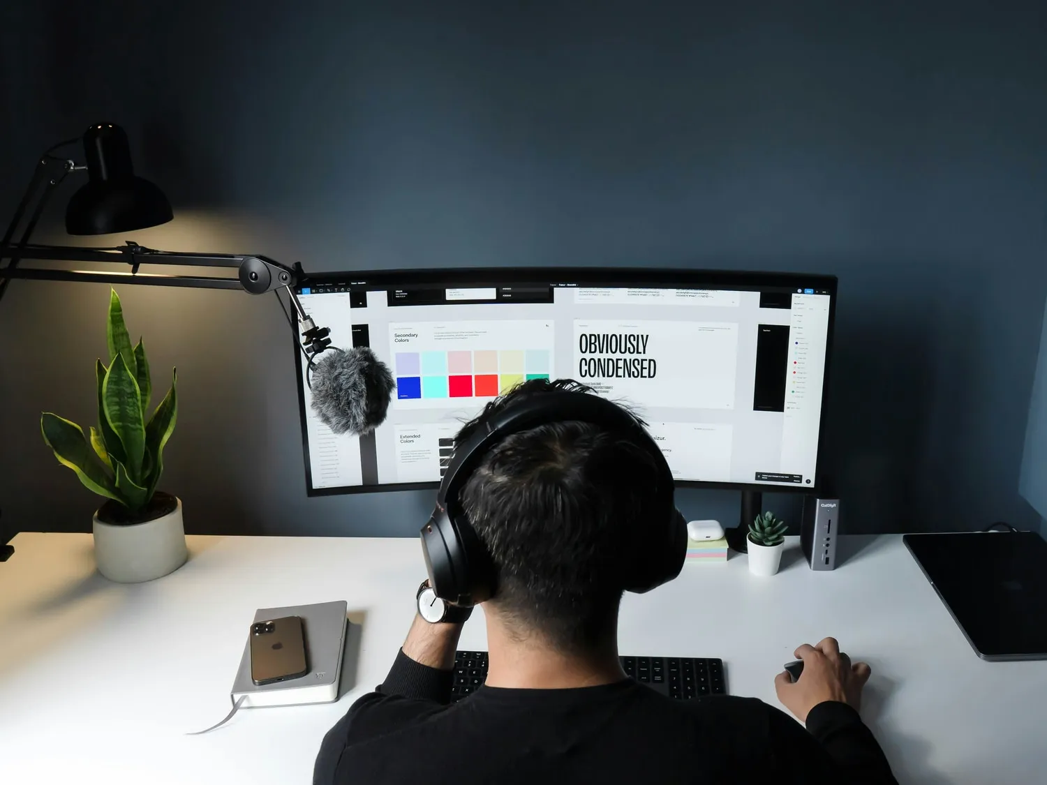 A designer at a desk, seen from behind, working on a brand style guide shown on a widescreen monitor.